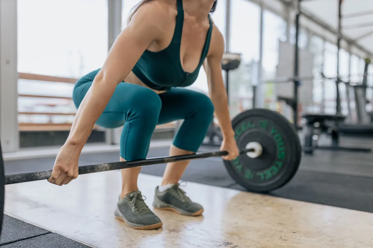 Vrouw in sportkleding pakt een barbell op voor een deadlift, met platte trainingsschoenen op een houten vloer