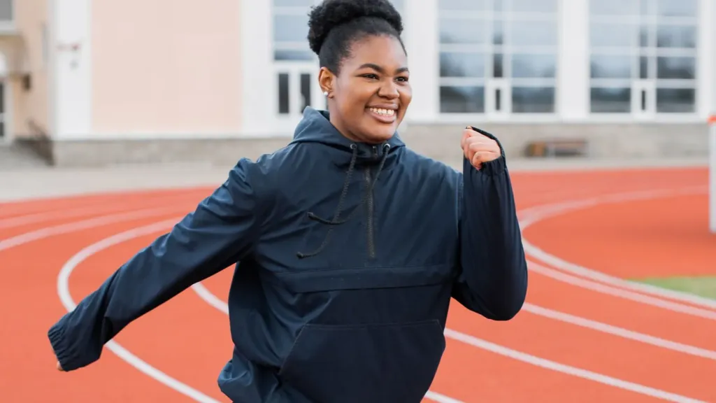 Lachende vrouw in een donkere trainingsjas rent met plezier over een rode atletiekbaan