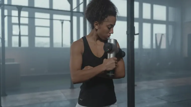 Vrouw in sporttop gebruikt een massage gun op haar bovenarm in een sportschool met grote ramen