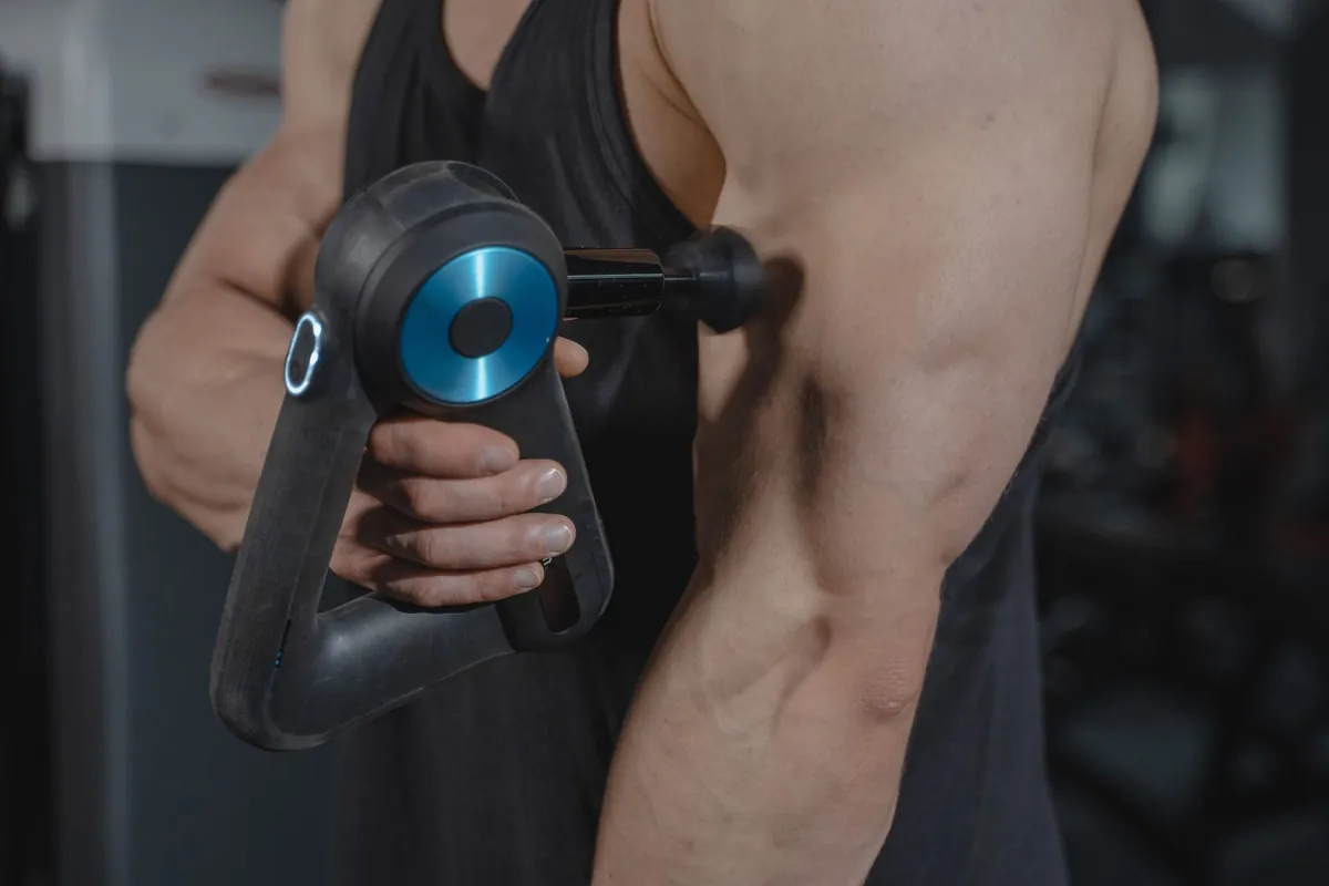Close-up van een zwart massagepistool met blauwe kop dat tegen de bovenarm van een sporter wordt gehouden