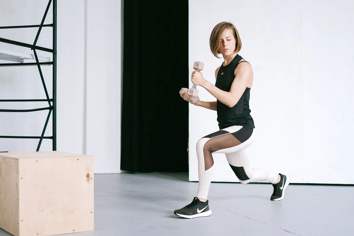 Vrouw doet een split squat met dumbbells naast een houten box in een lichte trainingsruimte