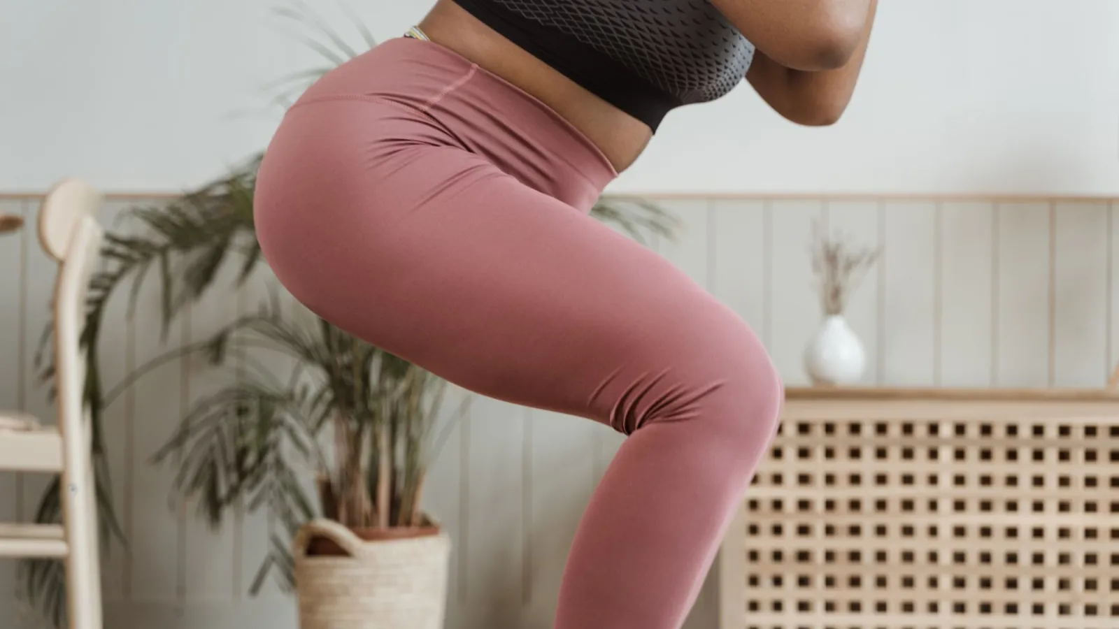 Vrouw in een roze sportlegging en zwart sporttopje doet een squat in een lichte woonkamer