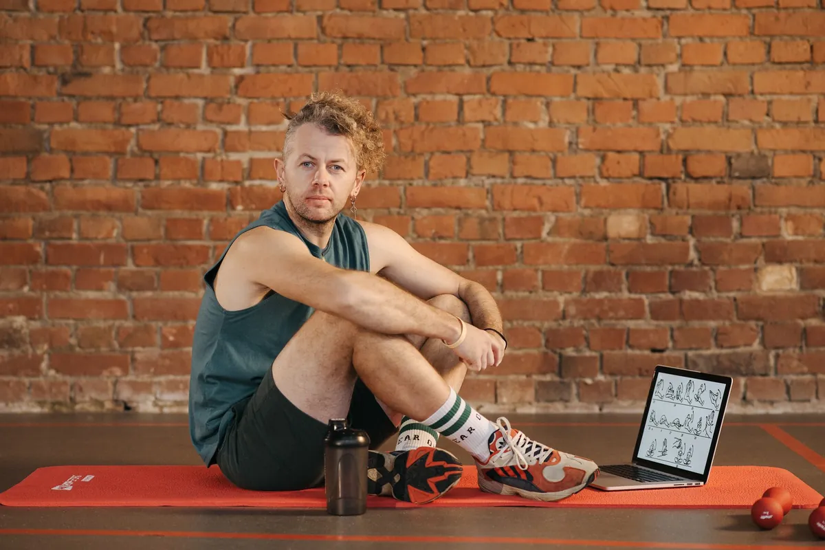 Man zit op een sportmat naast een laptop met een overzicht van oefeningen voor een thuistraining