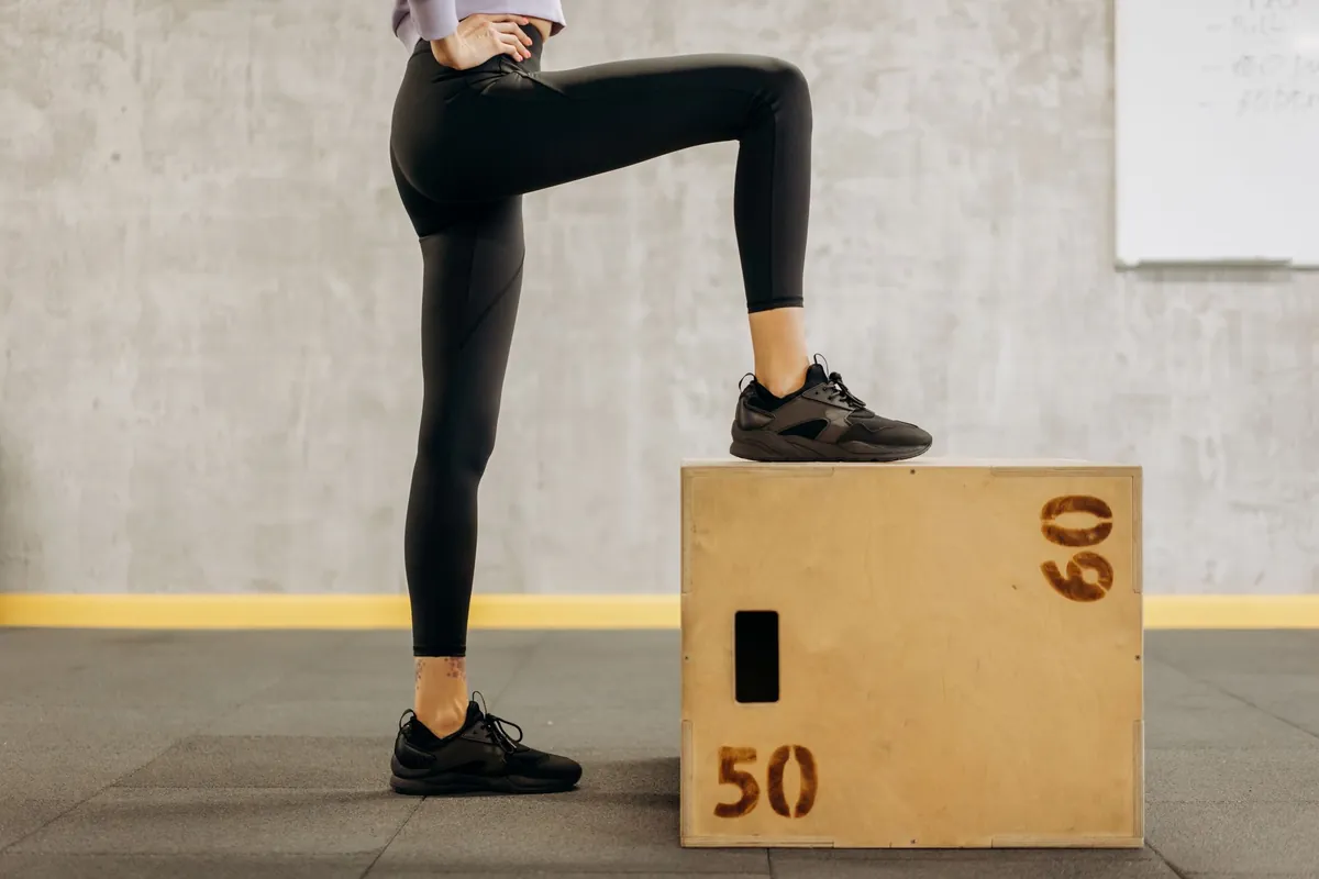 Vrouw in een zwarte high waist legging en sportschoenen zet een voet op een houten plyobox in de sportschool